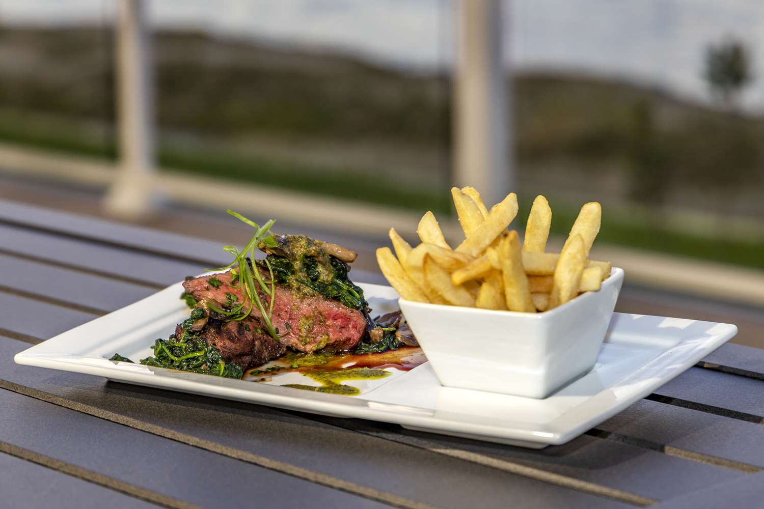 Sliced steak with greens and mushrooms, served on a rectangular white plate with a side dish of french fries on an outdoor table.