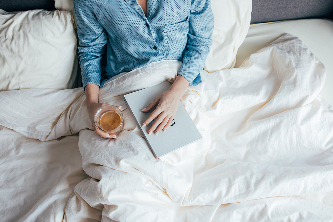 a person sitting in bed holding a book and a cup of coffee