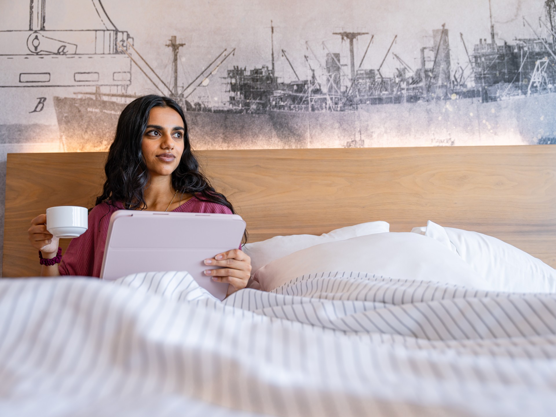 a woman sitting in bed holding a tablet and a cup of coffee