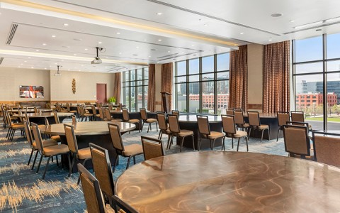 A spacious ballroom arranged with numerous large round tables and upholstered chairs over a blue patterned carpet.