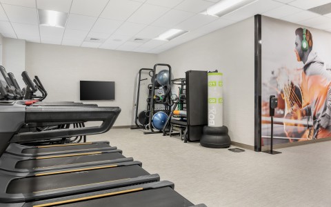 brightly lit hotel fitness center with a row of modern treadmills in the foreground. In the background, a storage rack holds exercise balls, mats, and other workout accessories next to a boxing bag.