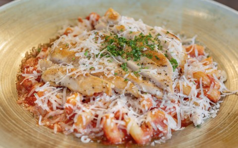 A close-up shot of a gourmet pasta dish served in a wide ceramic bowl. The dish features penne pasta tossed in red tomato sauce, topped with grilled, sliced chicken breast, a generous layer of shredded parmesan cheese, and a garnish of fresh chopped parsl