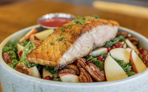 A close-up shot of a healthy salad bowl featuring a large, perfectly seared salmon filet sitting on a bed of fresh kale.