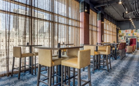 A lounge or dining space with high-top wooden tables and tan leather chairs arranged over a blue patterned carpet.