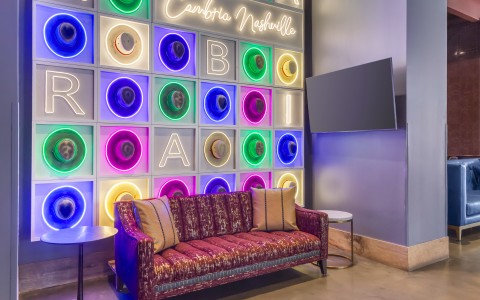 A vibrant lounge sitting area highlighting a large feature wall decorated with neon-lit squares, each framing a different colorful cowboy hat and spelling out "Cambria Nashville."