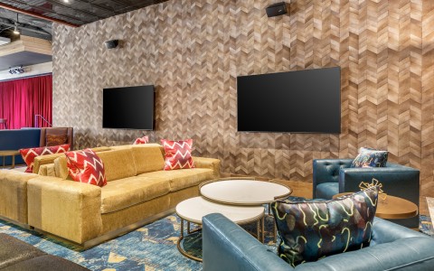 A chic lounge seating area featuring a large plush gold sofa with patterned red throw pillows. Two circular white coffee tables sit in the center, flanked by teal leather armchairs.