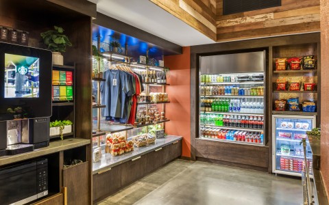 well-stocked hotel grab-and-go marketplace. The space features dark wood cabinets with open shelving displaying snacks, hanging T-shirts, souvenirs, and a coffee machine.