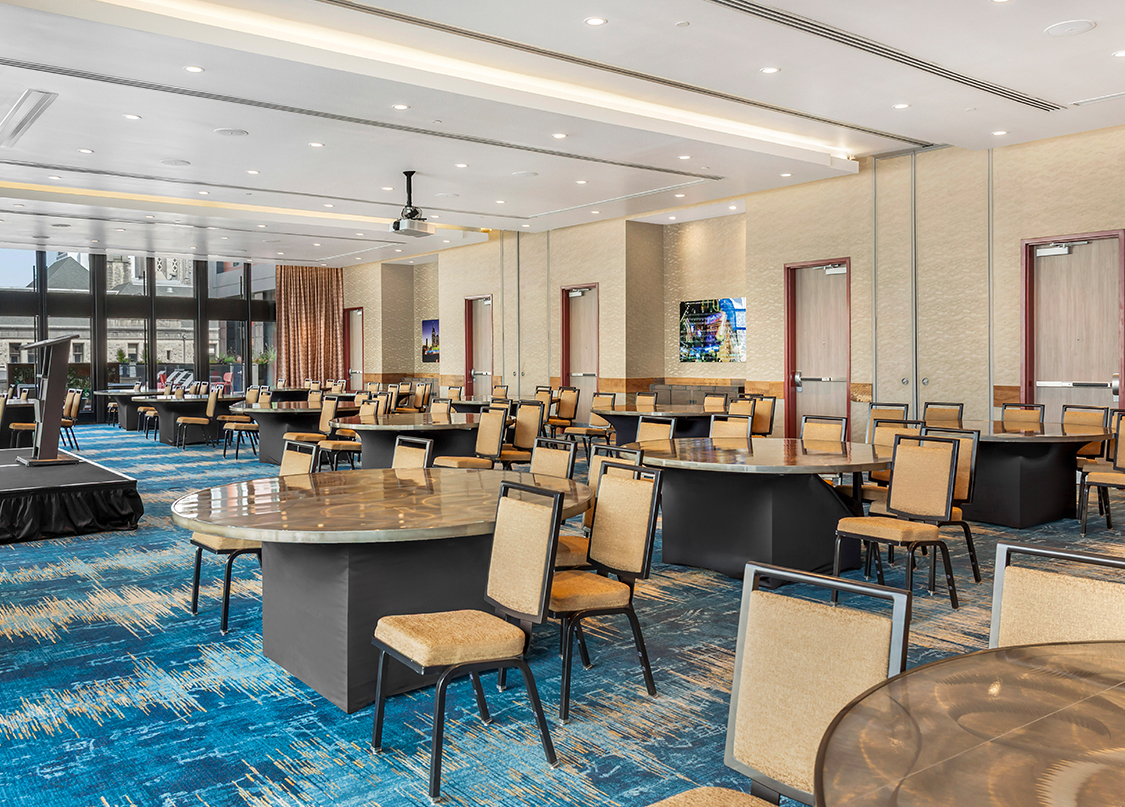 A spacious ballroom arranged with numerous large round tables and upholstered chairs over a blue patterned carpet.