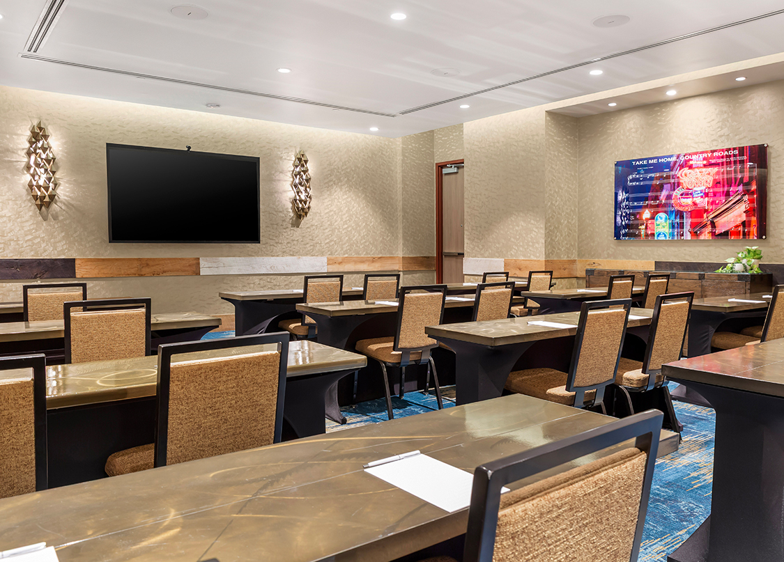 A classroom-style meeting room filled with several rows of long tables and tan upholstered chairs set up with paper and pens.