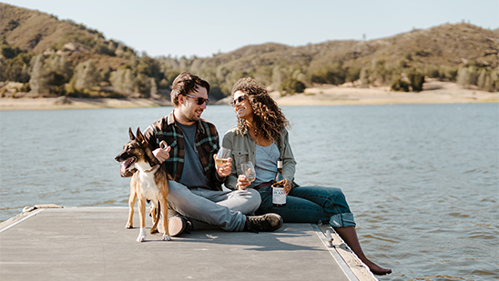 a man and woman sitting on a dock with a dog and a lake